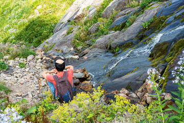 Rear view of a man in red with a backpack looking at the view of a small waterfall in the mountains while relaxing on a hike in summer