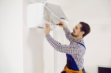 Male installation service worker or repairman installs or repairs air conditioner indoors. Young smiling man in workwear uses screwdriver to repair air conditioner that hangs on white wall in room.