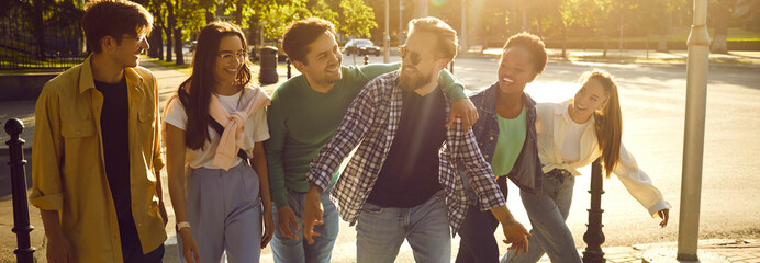 Happy guys and girls have fun walking streets of cities on sunny warm summer evening. Cheerful multiracial friends walking in row walking, talking and laughing. Friendship concept. Web banner.