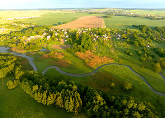 River near village, aerial view. Green fields and forest in rural. Country houses at river in countryside, drone view. Village with wooden house. Suburban house in rural on sunrise.