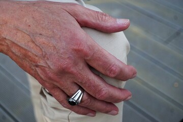 Schöne Hand von älterem Mann mit schwarzem Silberring auf Knie mit beiger Stoffhose bei Sonne am...