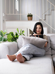 Attractive smiling young asian  woman relaxing on a leather couch at home, working on laptop computer.