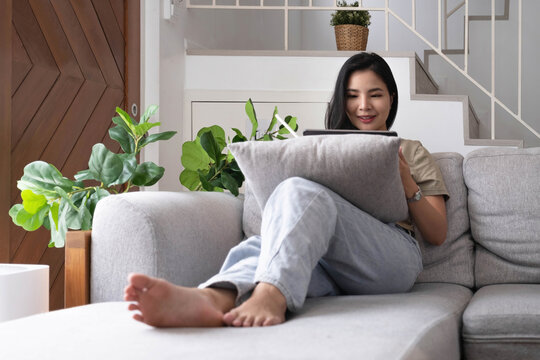 Attractive Smiling Young Asian  Woman Relaxing On A Leather Couch At Home, Working On Laptop Computer.