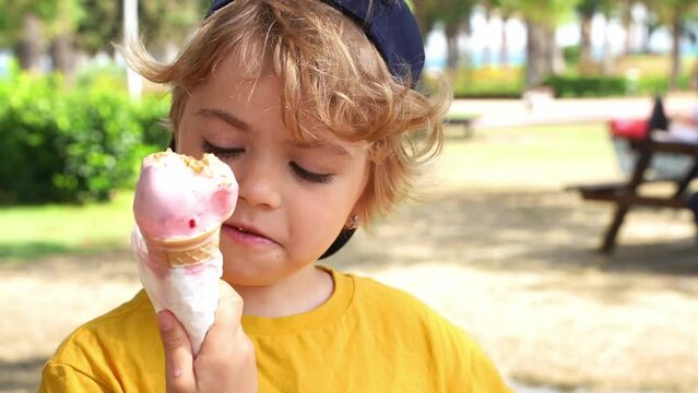 Preschooler child eating ice-cream in a waffle cone, licking sugar sweet dessert in a park during summer. Child wear yellow t-shirt and cap.