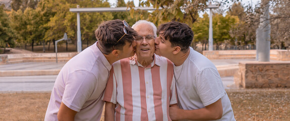 Dos nietos besando a su abuelo en el parque, con expresión cariñosa y felices. Fotografía en horizontal.