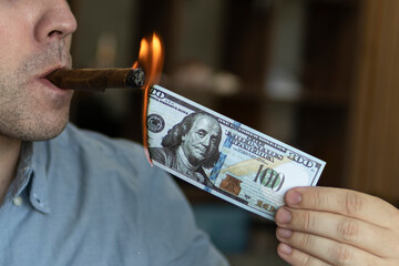 Young brutal man lighting cigar with 100 dollar bill as symbol of wealth and success