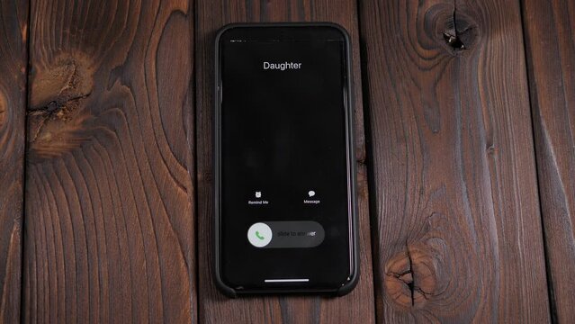 Close-up of an incoming call from a daughter on a mobile phone screen on a brown wooden background. Close-up of a woman answering her daughter's call.