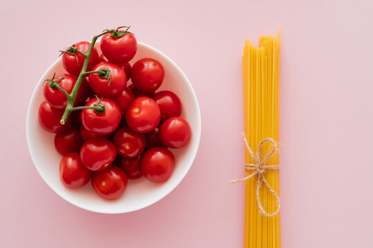 Top view of cherry tomatoes in bowl and uncooked spaghetti on pink surface.