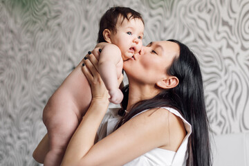 Portrait of happy young mother kissing tenderly little daughter in bedroom. Carefully hold naked baby in hands overhead. Selective focus, free copy space. Concept of maternal affection and childcare.