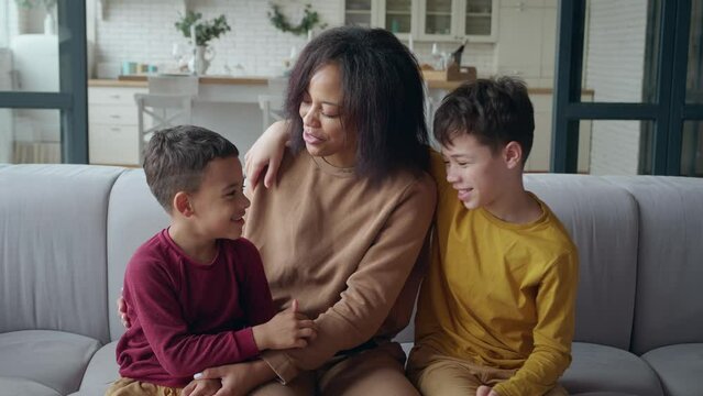 The boys sit on either side of their mom, swearing, arguing, and laughing together. A serene African American woman calms her sons, smiles and rejoices with them sitting on the couch in the living rom