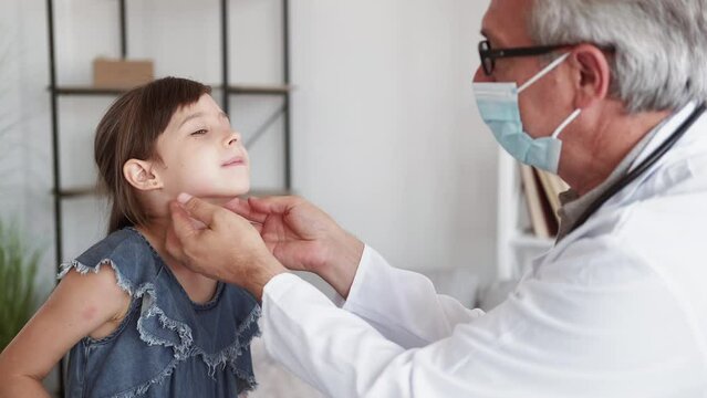 Pediatrician visit. Kid checkup. Respiratory disease. Child medical care. Middle-aged male doctor in protective face mask examining ill little girl patient sore throat.