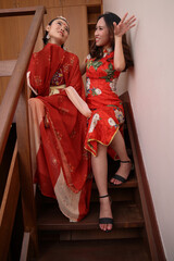 two young attractive Asian woman wearing tadeonal Chinese red hanfu long skirt scarf and cheongsam costume dress sit on indoor wooden stair pose talk