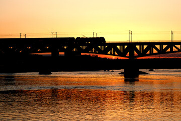 The train riding over the bridge over the river at sunset.