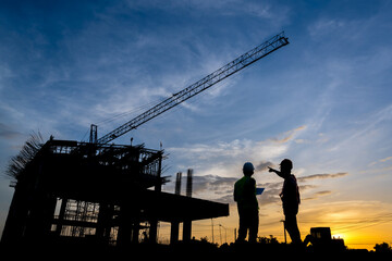 Silhouette of a male engineer standing at a construction site in the evening sunset. A team of construction engineers talks to managers and construction workers at an industrial construction project.