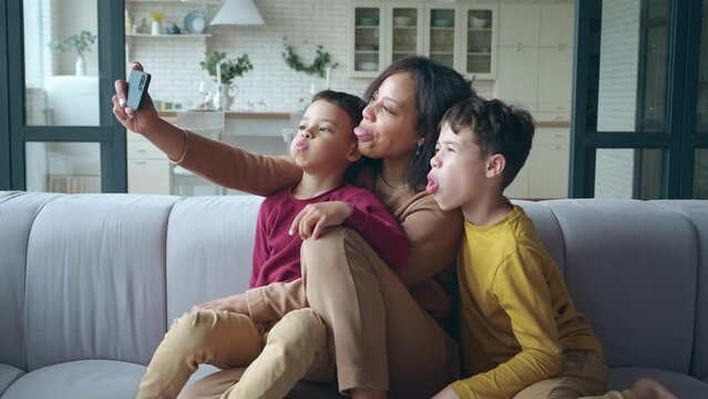 A Cheerful, Joyful And Playful Mixed Race Woman Sits On The Sofa In The Living Room, Tenderly Hugs Her Sons, Frowns And Shows Her Tongues In Front Of A Mobile Phone Camera In An Outstretched Hand.