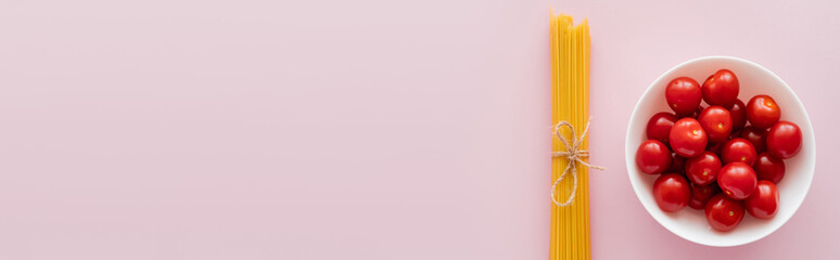 Top view of raw spaghetti and cherry tomatoes in bowl on pink surface, banner.