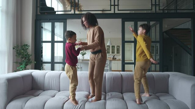 Happy African American woman enjoying family time with her two sons, jumping and dancing with them in turns on the couch. Happy family of a mom and two funny and joyful boys chilling together at home