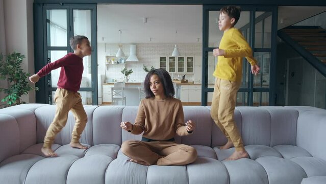 Boys make noise while jumping on the couch in the side of their exhausted mother. Tired African American woman shakes her head, closes her eyes, begins to meditate in lotus position. Mom performs yoga