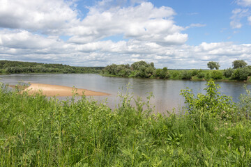 Summer rural landscape. Green meadow with tall grass, blue sky with white clouds, river on a sunny day.