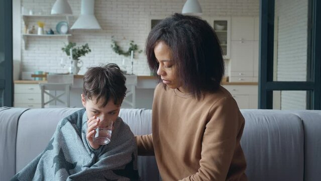 A worried and sad young mother gently wraps her sick son with a warm blanket and gives him a glass of water to drink. Boy with signs of infectious disease and fever leaning on caring mother.