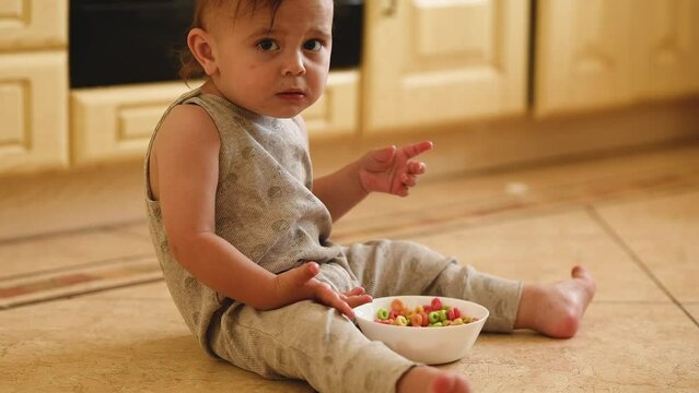 a one-year-old baby is sitting at home on the kitchen floor and eating multicolored cereal flakes from a white plate. The child eats a dry breakfast