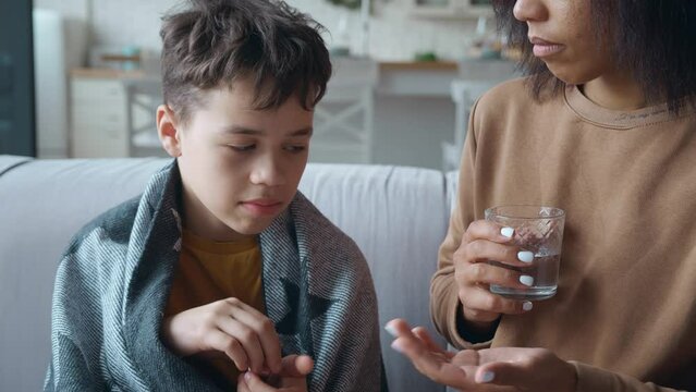 Close-up portrait of a mixed race boy, wrapped in a blanket, sitting next to his mother, taking medicine and a glass of water to treat his illness. The child caught a cold and is being treated at home