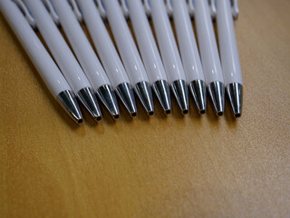 Group of ten white plastic with metal tips ballpoint pens lying flat on beige lacquered wooden table top.