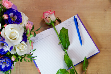Flatlay. Notepad, diary with a pen on a wooden table near a bouquet of multi-colored roses. Workspace with diary and pink white flower on wooden background. Top view
