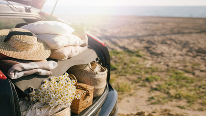 Open trunk of car with picnic items: straw hat, basket with food and flowers with summer beach on the background. © polinaloves