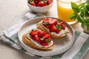 Homemade bruschettas with strawberries, cream cheese and basil in white plate on light background. Summer appetizer. Selective focus.