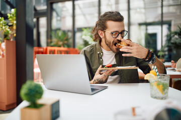 Young businessman eats sandwich while working on laptop in cafe.