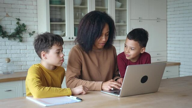 Smiling African American mother and kids sons having fun with computer sitting at the kitchen table. Happy mixed race mom with childs boys using laptop, watching cartoons, making video call, shopping