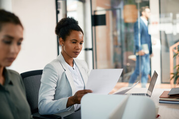Black businesswoman going through paperwork while working on laptop in office.