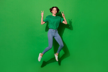 Fototapeta na wymiar Photo of funny adorable woman dressed summer t-shirt jumping high showing v-signs isolated green color background