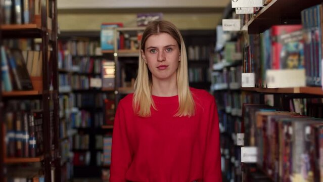 Caucasian blonde woman in a red sweater is standing in the library among the shelves with books, looking around and looking into camera, portrait