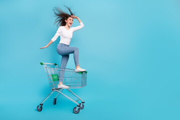 Photo of funky adorable woman wear white shirt riding shopping tray looking far away empty space isolated blue color background