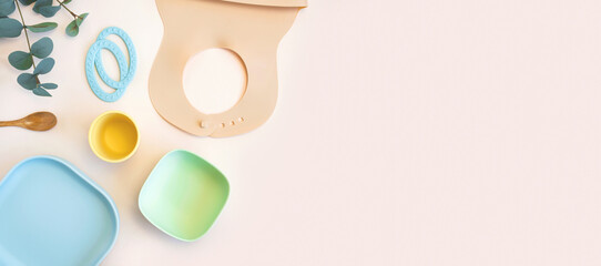 Baby accessories and tableware for food on beige background. First feeding for kids concept. Flat lay, Top View, Space for text. Silicone dishware, bib, cup on neutral backdrop. Serving baby food