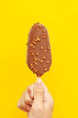 A hand holds an ice cream with nuts and chocolate icing on a yellow background. Vertical photo. Close-up.