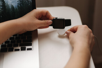 Female hand put smart watch on wireless charging with on-screen charging indicator. Work place near at the laptop