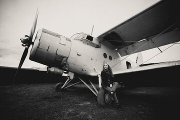 Art work. A beautiful girl is sitting on the wing of an airplane, she is bored. Getting ready to fly. The plane stands on the grass in a clearing. The photograph is black and white.