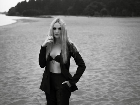 Art work. A black and white photo of a beautiful stylish girl, with blond hair, in a black classic suit and black lingerie, who is standing on the sand, on the beach and smoking.