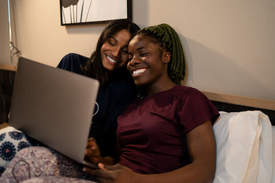 Happy diverse female friends watching video on laptop on bed