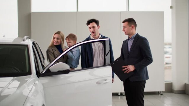 young man and woman with a little boy in their arms are choosing a new car for their family and talking to a male salesman in a auto sales center