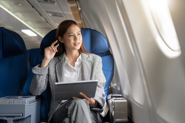Successful beautiful young asian business woman sits in airplane cabinplane and works on digital...