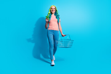 Full size photo of young charming woman go on shopping buy groceries hold smartphone isolated on blue color background