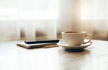 Close up view, white coffee cup with smartphone, notebook and pen on table, after get up in the morning on holiday with breakfast, vintage color tone and soft light from window on background