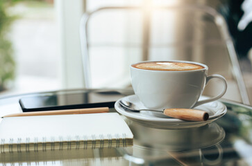 Close up view, latte coffee in white cup with smartphone, notebook and pen on table, after get up in the morning on holiday with breakfast