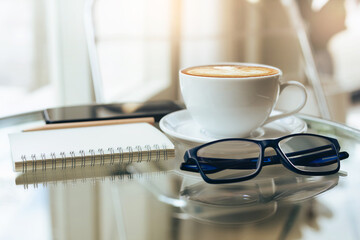 Close up view, eyeglasses and note book with latte coffee in white cup and mobile phone on table, relax in cafe after work and soft light from window on background