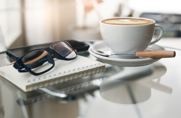 Close up view, eyeglasses on note book with latte coffee in white cup and mobile phone on table, relax in cafe after work and soft light from window on background