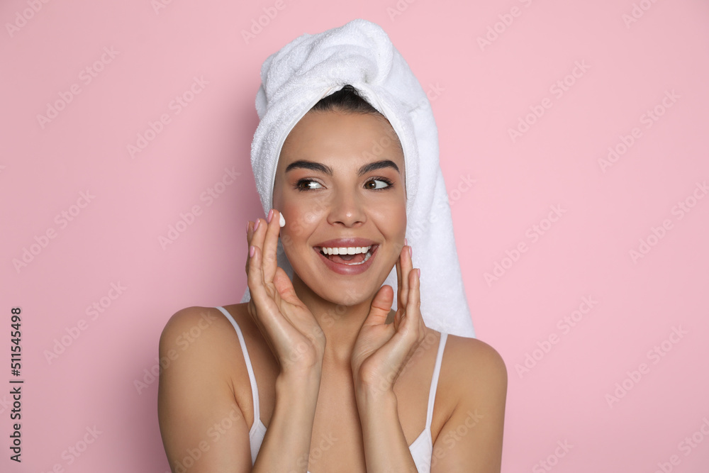 Wall mural beautiful young woman with towel applying cream on face against pink background - Wall murals